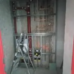 Wnętrze w trakcie montażu instalacji wodnej: widoczne rury miedziane, metalowa konstrukcja wsporcza, rozdzielacze z zaworami i drabina aluminiowa. Ściany surowe, betonowe.