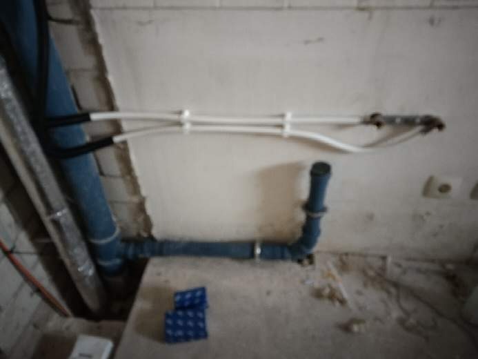 Fragment instalacji hydraulicznej z rurami PCV w trakcie montażu, widoczne białe rury mocowane do ściany i niebieskie rury podłączone do odpływu, w tle surowa ściana.