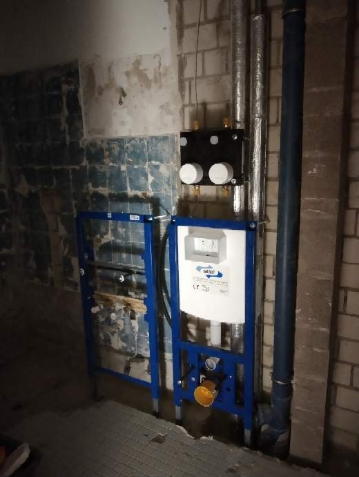 Stelaż podtynkowy WC zamontowany w surowym wnętrzu, widoczne rury instalacyjne i fragmenty starej glazury na ścianach.