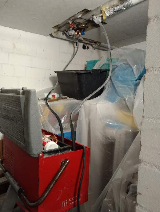 Instalacja hydrauliczna pod stropem z widocznymi rurami miedzianymi i zaworami, czarne rury podłączone do czerwonej maszyny z szarym wiekiem, całość w pomieszczeniu z surowymi ścianami...