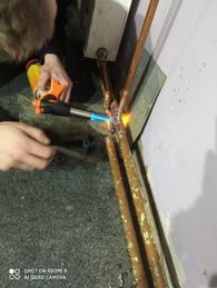 Miedziane rury lutowane palnikiem gazowym przy ścianie, widoczne narzędzia i ręce pracownika w trakcie łączenia instalacji.