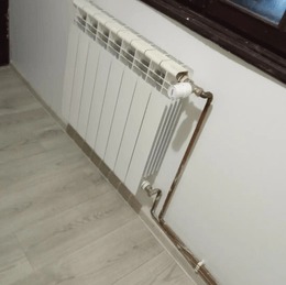 Biały, aluminiowy grzejnik panelowy zamontowany przy ścianie, z widocznymi miedzianymi rurami doprowadzającymi, termostat na górze, widoczne fragmenty podłogi i okna.