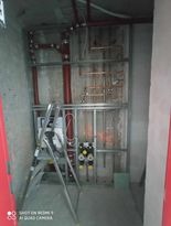 Wnętrze w trakcie montażu instalacji wodnej: widoczne rury miedziane, metalowa konstrukcja wsporcza, rozdzielacze z zaworami i drabina aluminiowa. Ściany surowe, betonowe.