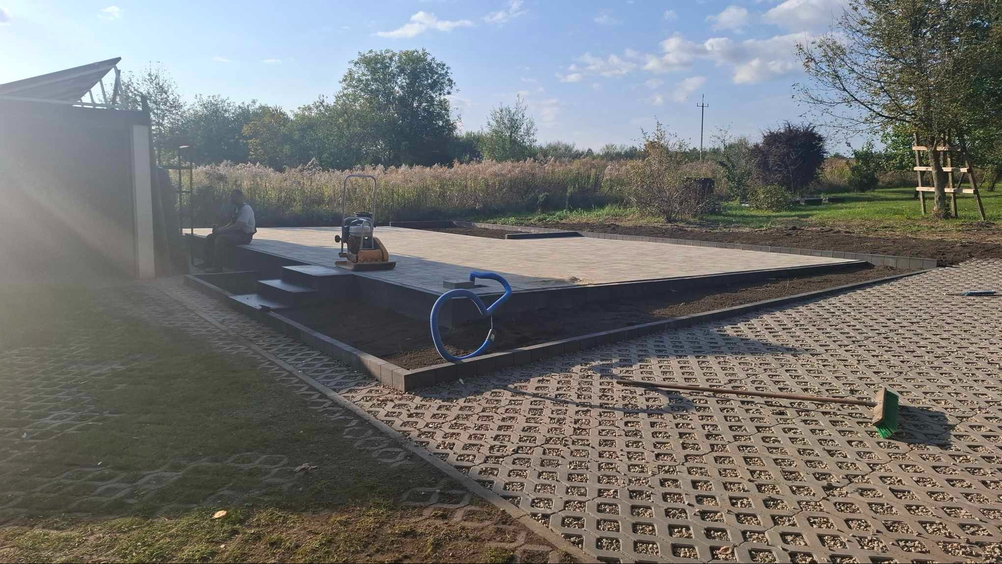 Nierówna nawierzchnia z ażurowych płyt betonowych przechodzi w świeżo ułożony plac z szarej kostki, z ubijarką wibracyjną i niebieskim wężem w tle, pracownik siedzący z boku.