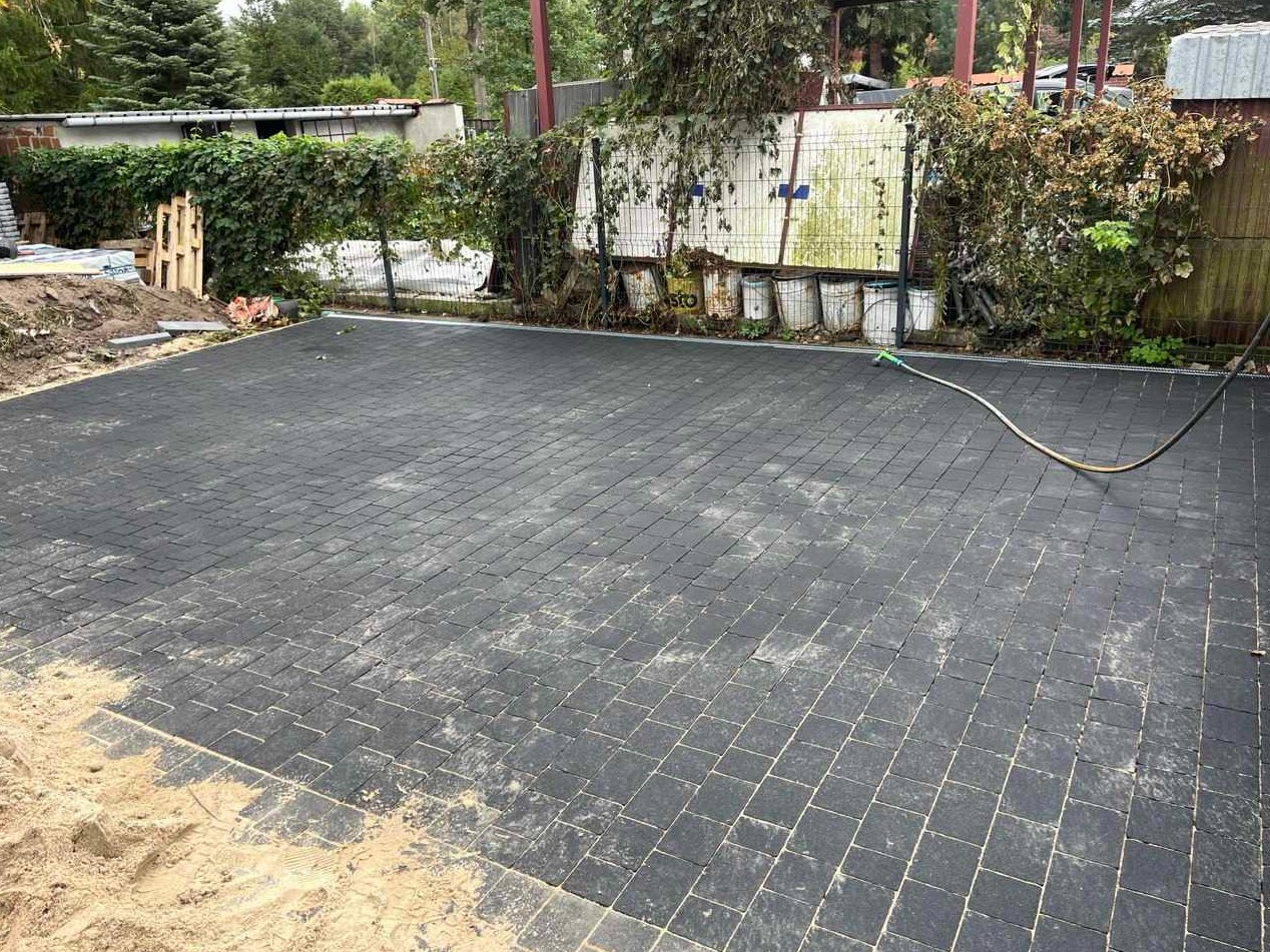 Ukończona powierzchnia z czarnej kostki brukowej ułożonej w regularny wzór, z widocznym fragmentem piasku na krawędzi i wężem ogrodowym leżącym na powierzchni, otoczona zielenią i ogrodzeniem.