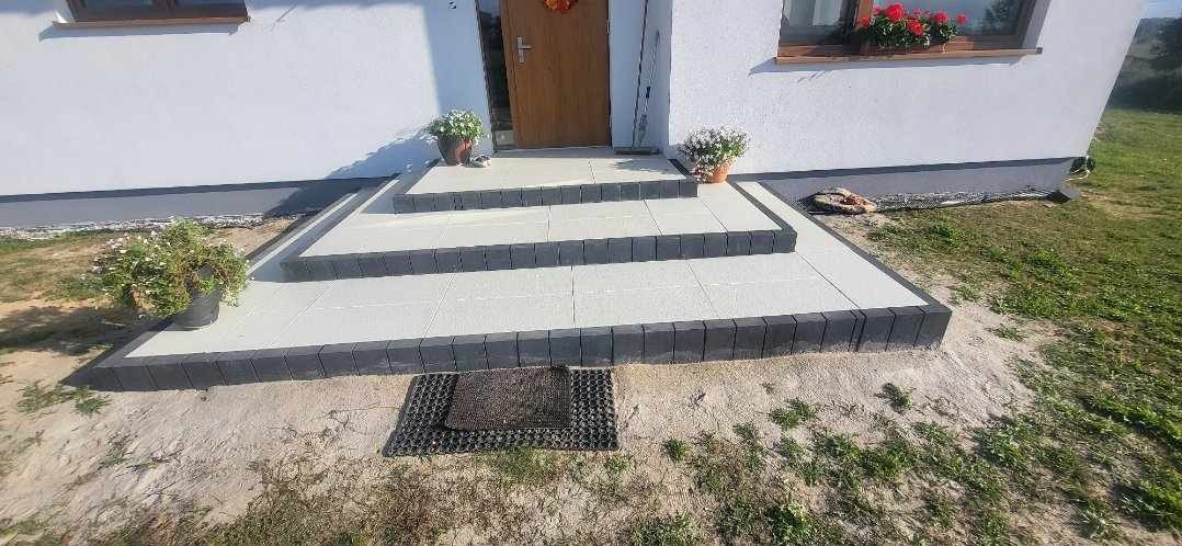 Schody wejściowe do domu wyłożone jasnymi płytami z ciemną obwódką, z doniczkami z kwiatami na każdym stopniu, widok z dołu na dom z białymi ścianami.