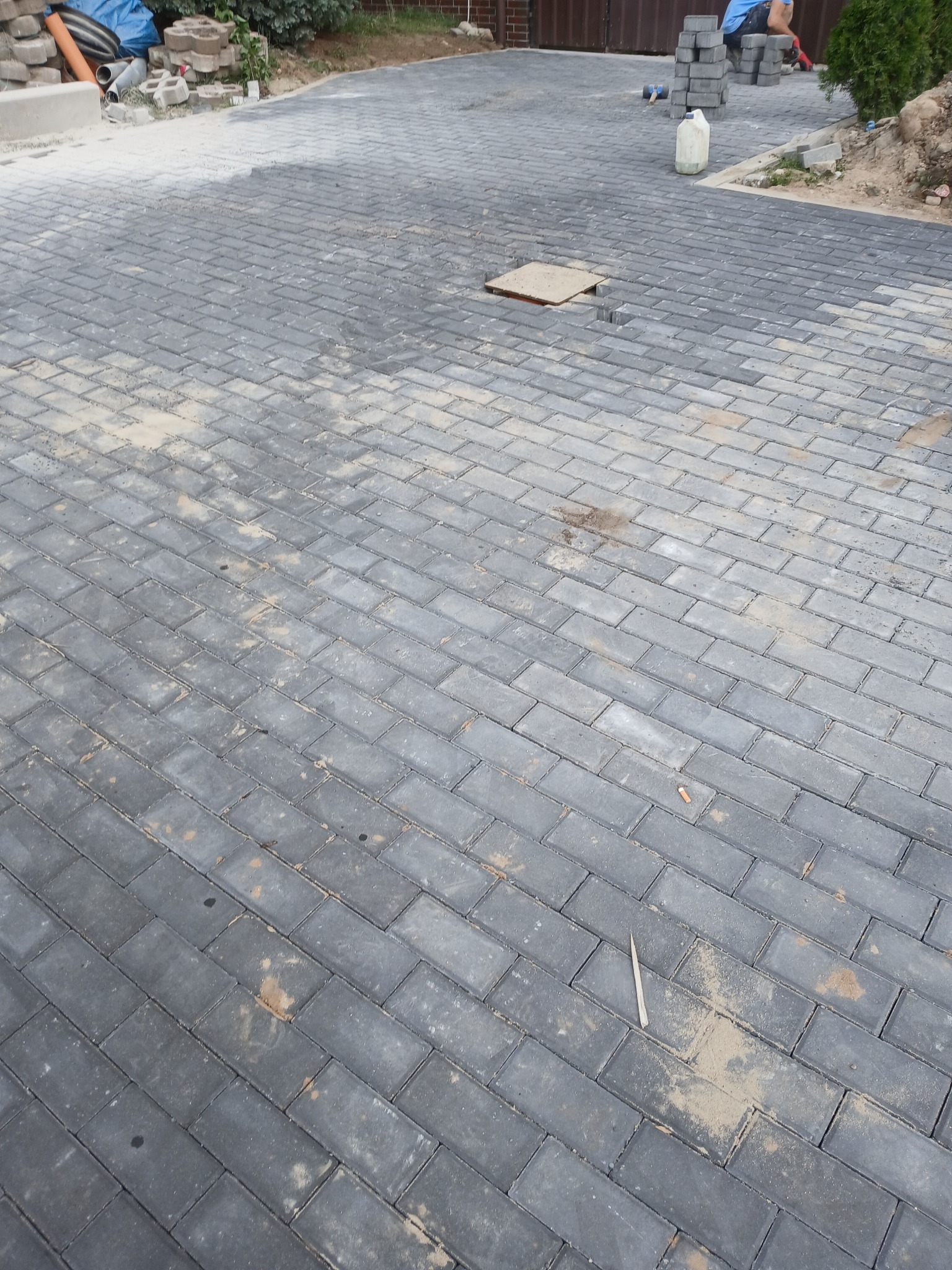 Częściowo ułożona, szara kostka brukowa w kształcie prostokątów, z widocznymi śladami piasku i narzędziem do fugowania, fragmentem niezakończonej studzienki rewizyjnej oraz stertą kostki...