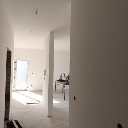 MiniBud remonty i wykończenia Roman Havryliuk - Wnętrze w trakcie remontu: świeżo pomalowane ściany na biało, widoczny sprzęt do cięcia płytek na stole, narzędzia malarskie w kuwecie, surowy beton na podłodze i przygotowane otwory na gniazdka.