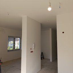MiniBud remonty i wykończenia Roman Havryliuk - Wnętrze w stanie deweloperskim po malowaniu, z widocznymi puszkami elektrycznymi, wiszącą żarówką i drabiną w tle.