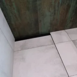 Połączenie płytek podłogowych w odcieniach szarości z płytkami ściennymi imitującymi rdzę, oddzielone czarną listwą wykończeniową.