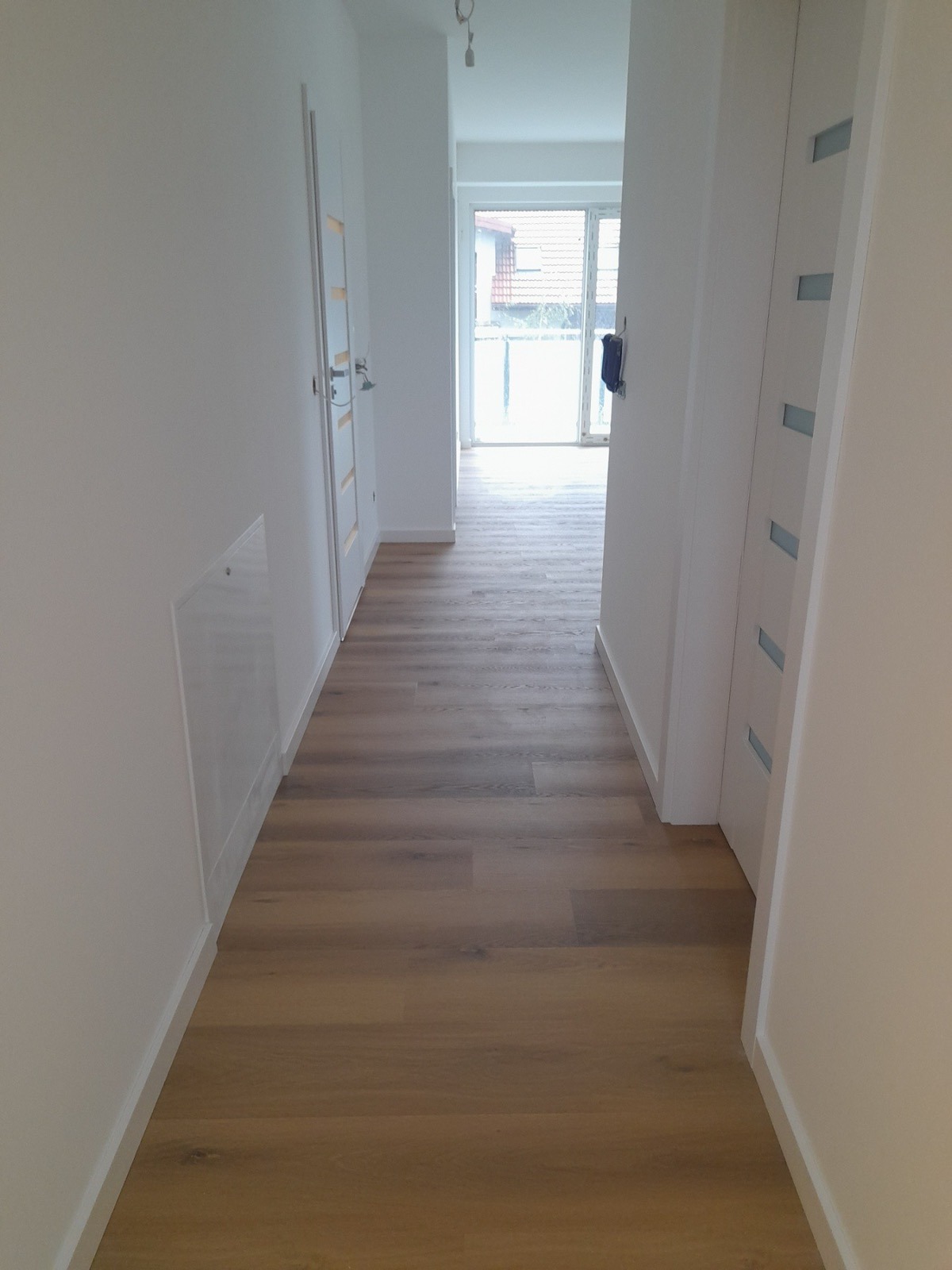 Korytarz z panelami podłogowymi w odcieniach brązu, białe ściany i drzwi z przeszkleniami, widok na jasne pomieszczenie z balkonem w tle.