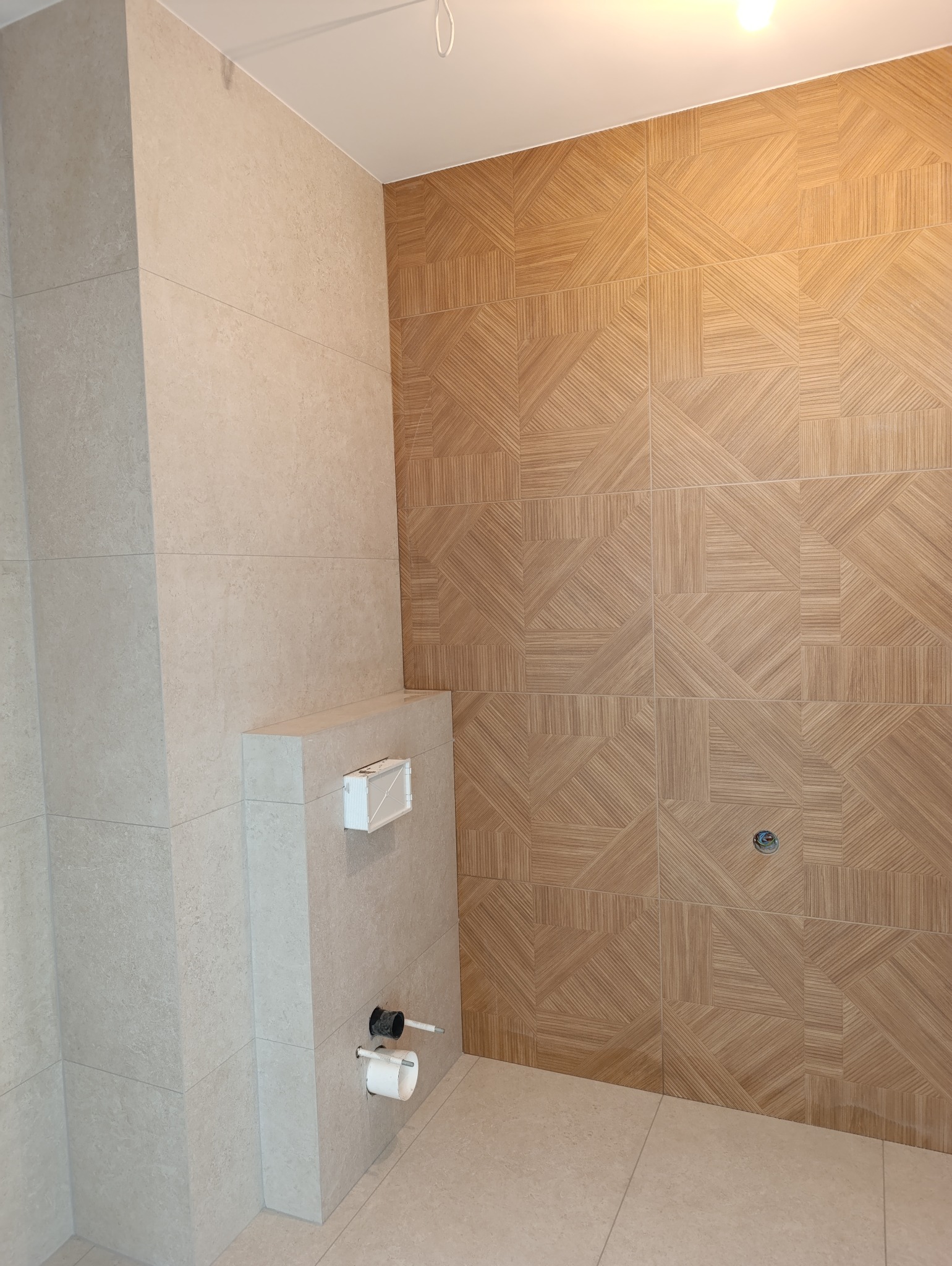 Nowoczesna łazienka z beżowymi i drewnopodobnymi płytkami, widoczny zabudowany stelaż podtynkowy WC i fragment instalacji wodno-kanalizacyjnej.