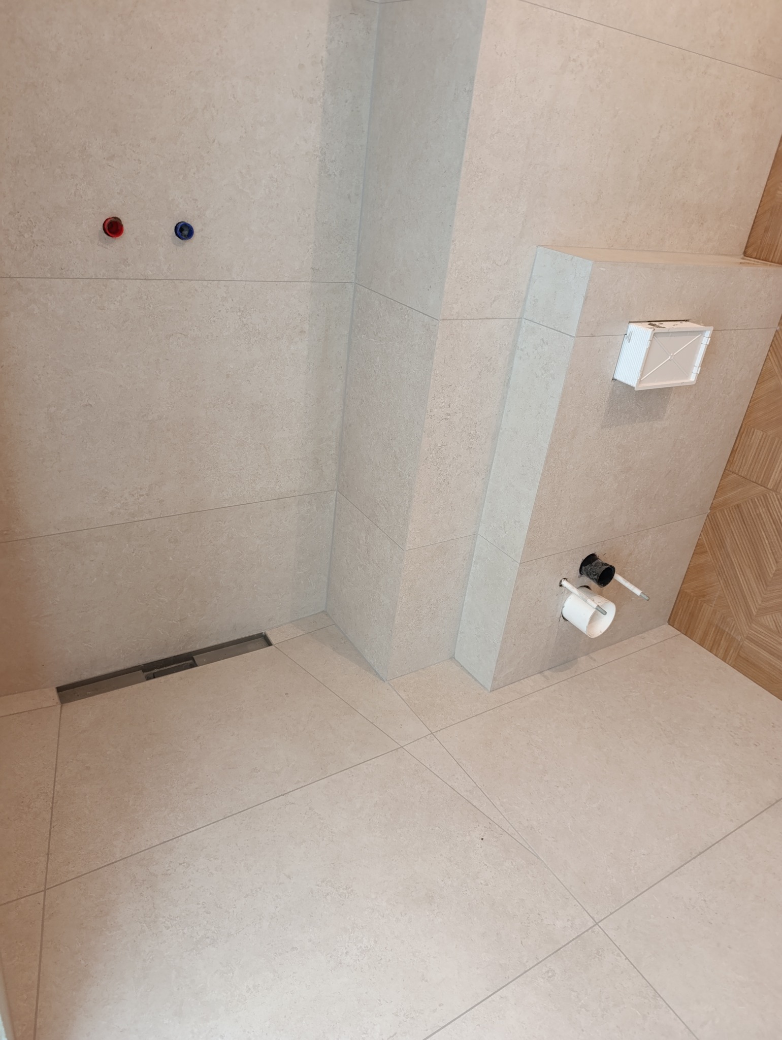 Wykończona łazienka z beżowymi płytkami, widoczny odpływ liniowy, podtynkowa spłuczka WC i wyprowadzenia instalacji wodnej z zaślepkami.