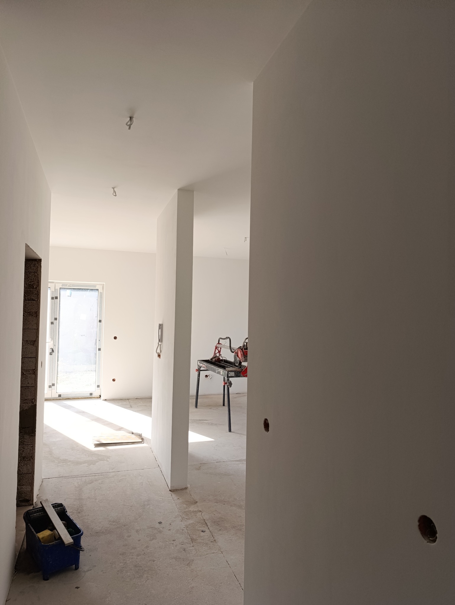 Wnętrze w trakcie remontu: świeżo pomalowane ściany na biało, widoczny sprzęt do cięcia płytek na stole, narzędzia malarskie w kuwecie, surowy beton na podłodze i przygotowane otwory na gniazdka.
