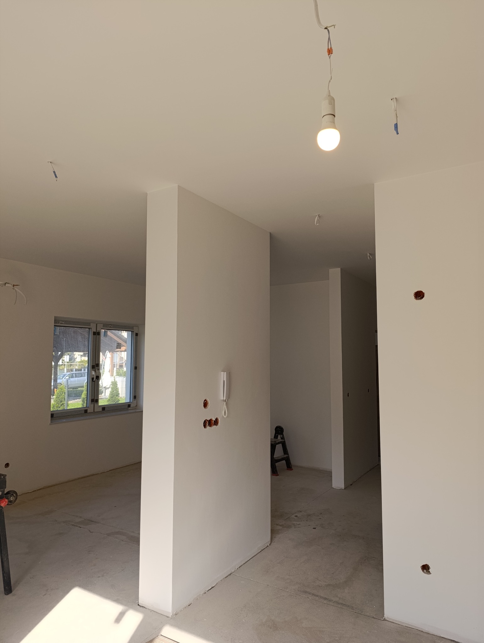 Wnętrze w stanie deweloperskim po malowaniu, z widocznymi puszkami elektrycznymi, wiszącą żarówką i drabiną w tle.
