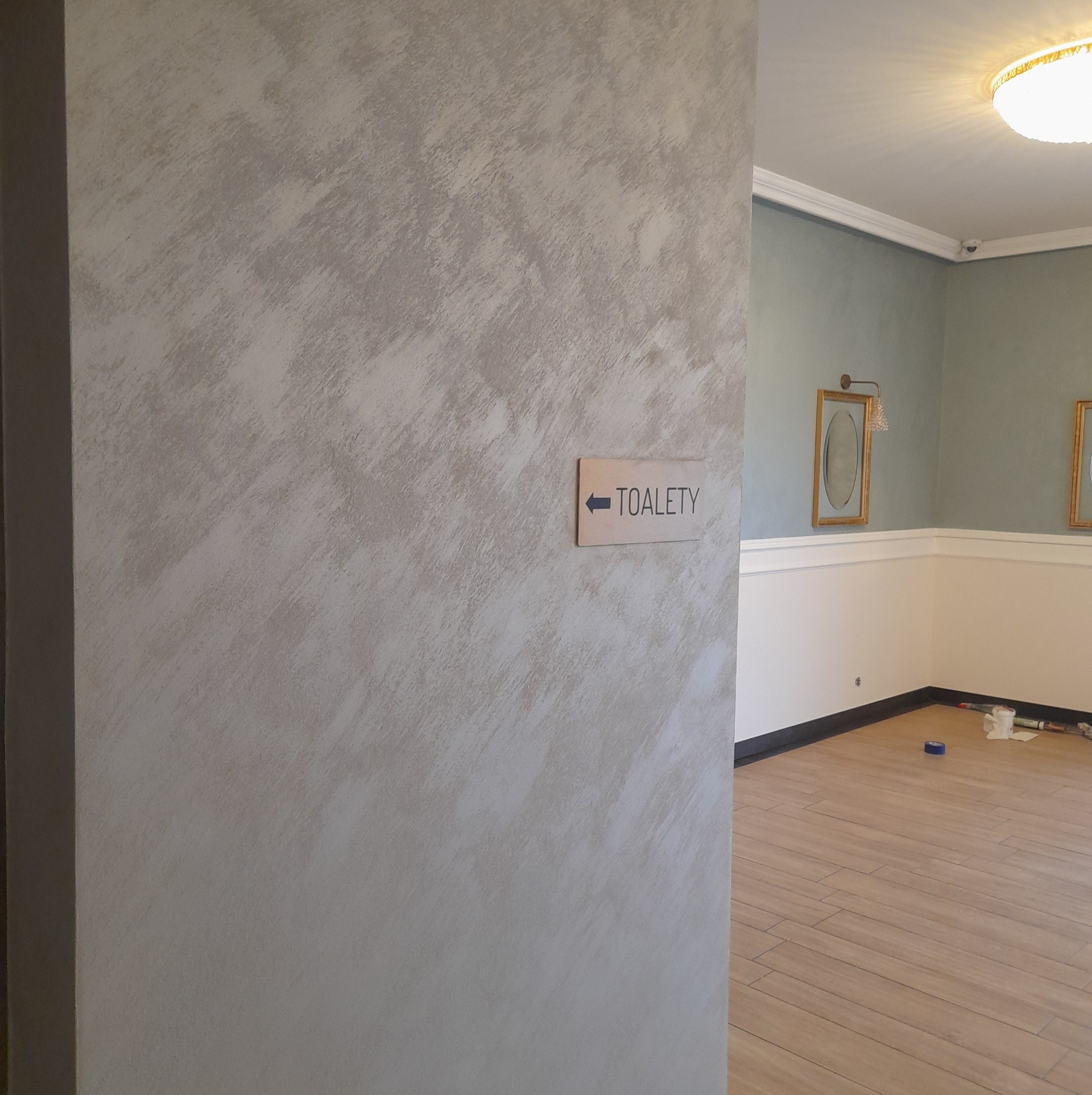 Ściana z dekoracyjną, beżowo-szarą fakturą, wskazująca kierunek do toalet, widoczne fragmenty odnowionego wnętrza z listwami przypodłogowymi i drewnopodobną podłogą.