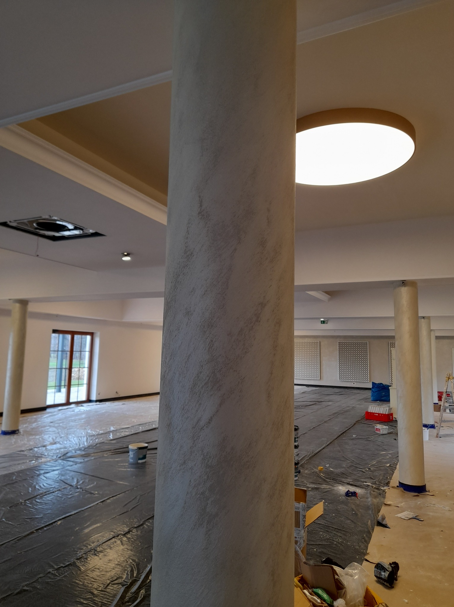 Wnętrze w trakcie prac wykończeniowych: kolumna z dekoracyjną strukturą, zabezpieczona podłoga folią, widoczne narzędzia malarskie i puszki z farbą.