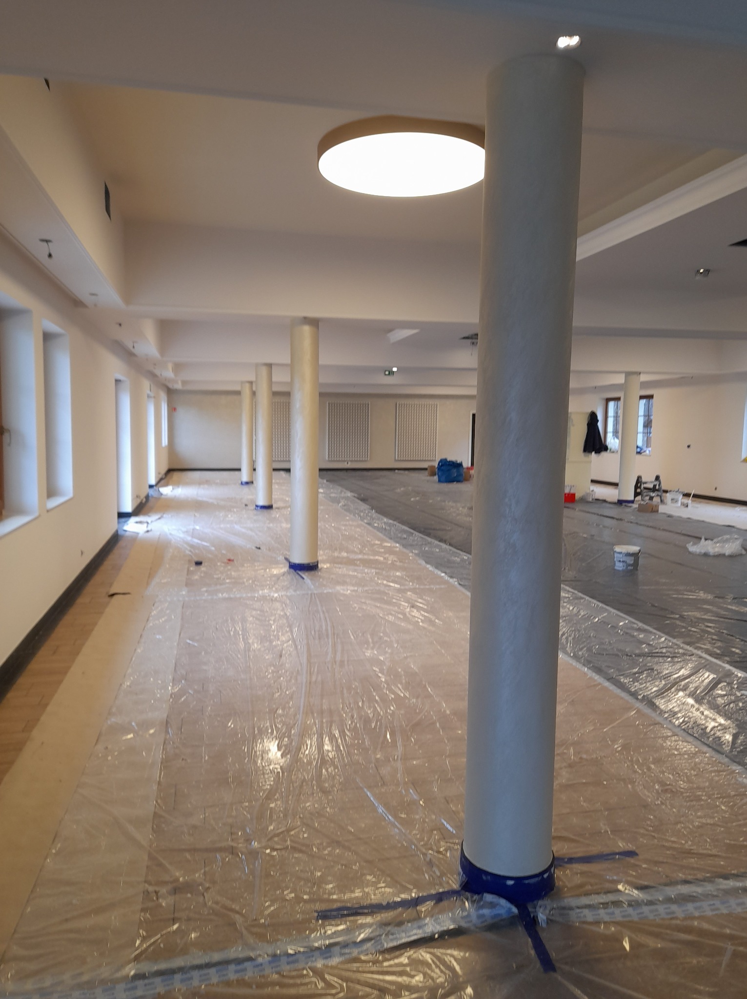 Przygotowane do malowania, zabezpieczone folią podłogi i kolumny w dużym pomieszczeniu biurowym, widoczne wiadra z farbą w tle.
