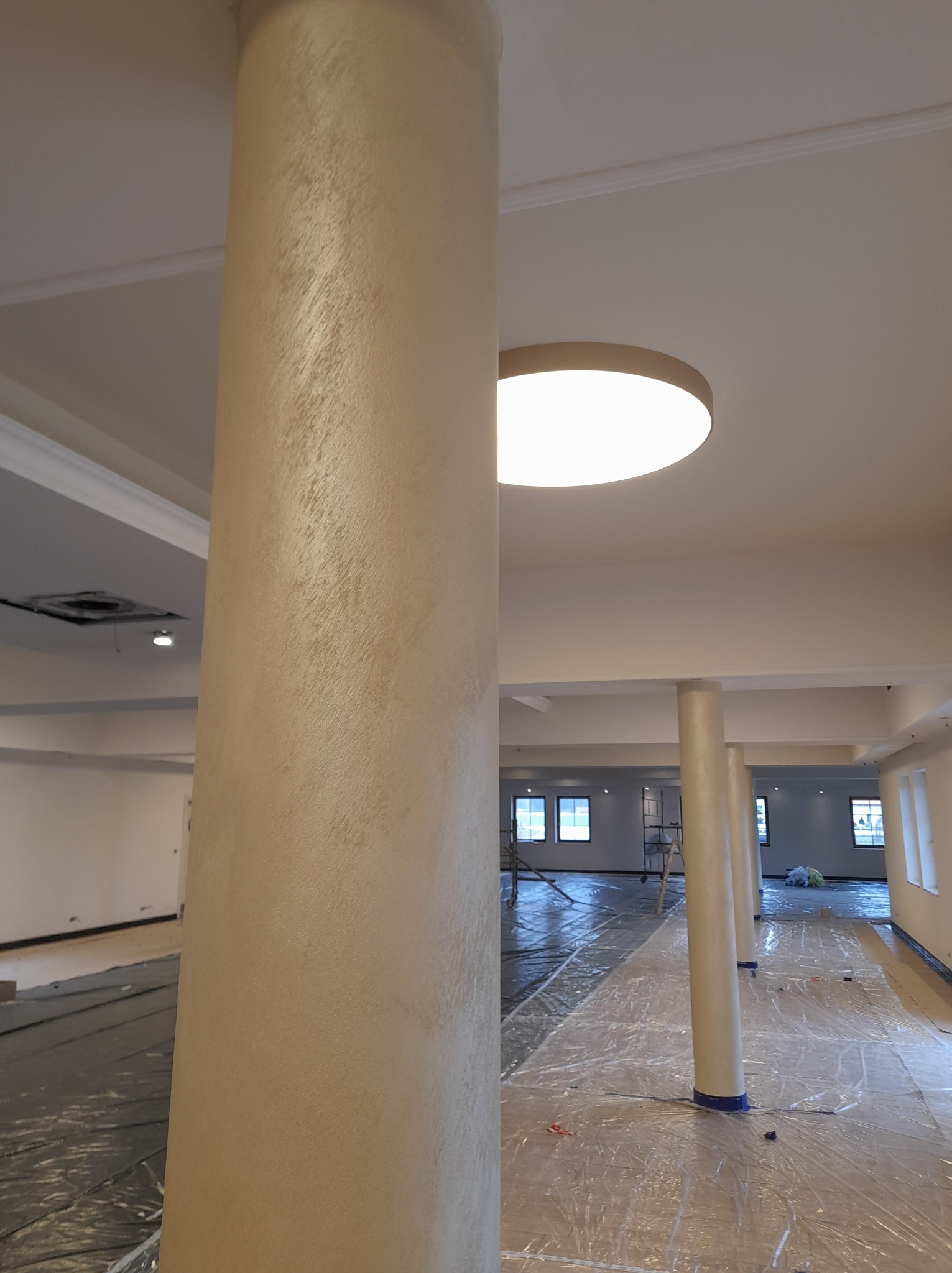 Wykończony filar w odcieniach beżu z dekoracyjną fakturą, widoczne zabezpieczone folią podłoże i sufit z oświetleniem w trakcie prac wykończeniowych w przestronnym wnętrzu.