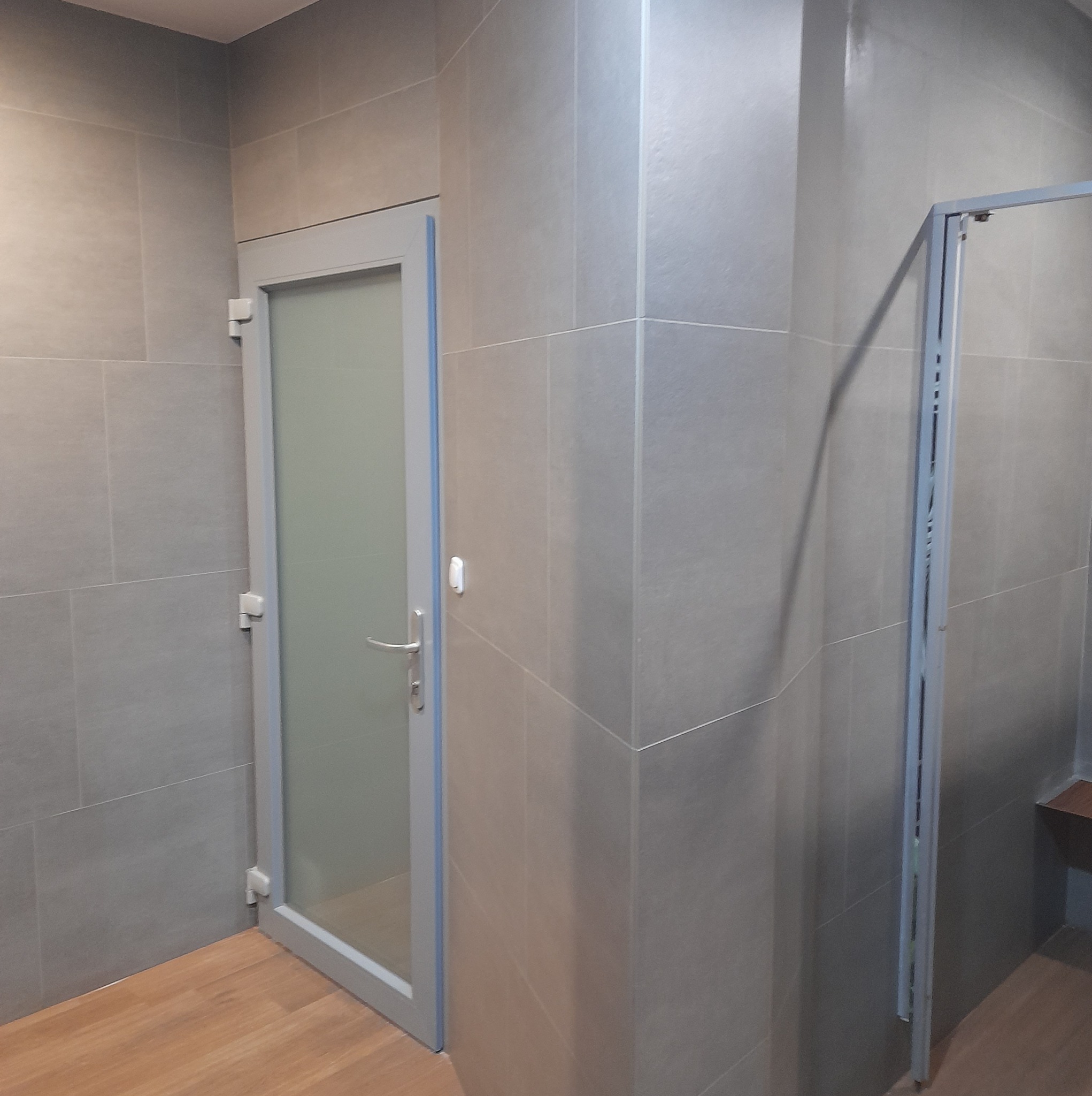 Wykończona łazienka z szarymi płytkami, kabiną prysznicową z metalową ramą i drewnopodobną podłogą, widok na drzwi z mleczną szybą.