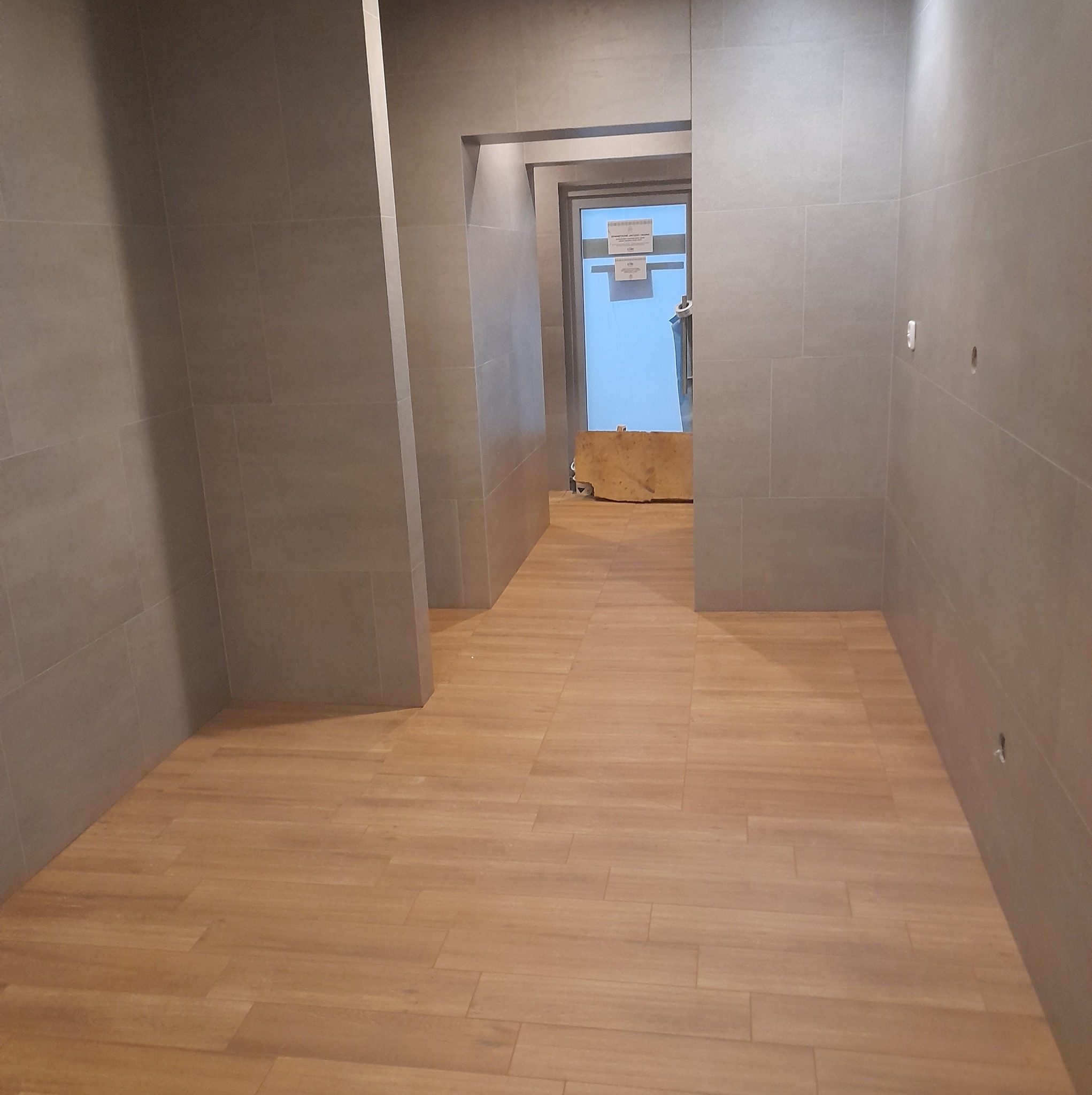 Wnętrze korytarza wykończonego szarymi płytkami ściennymi i panelami podłogowymi w odcieniach drewna, widoczne otwory w ścianie przygotowane pod gniazdka elektryczne.