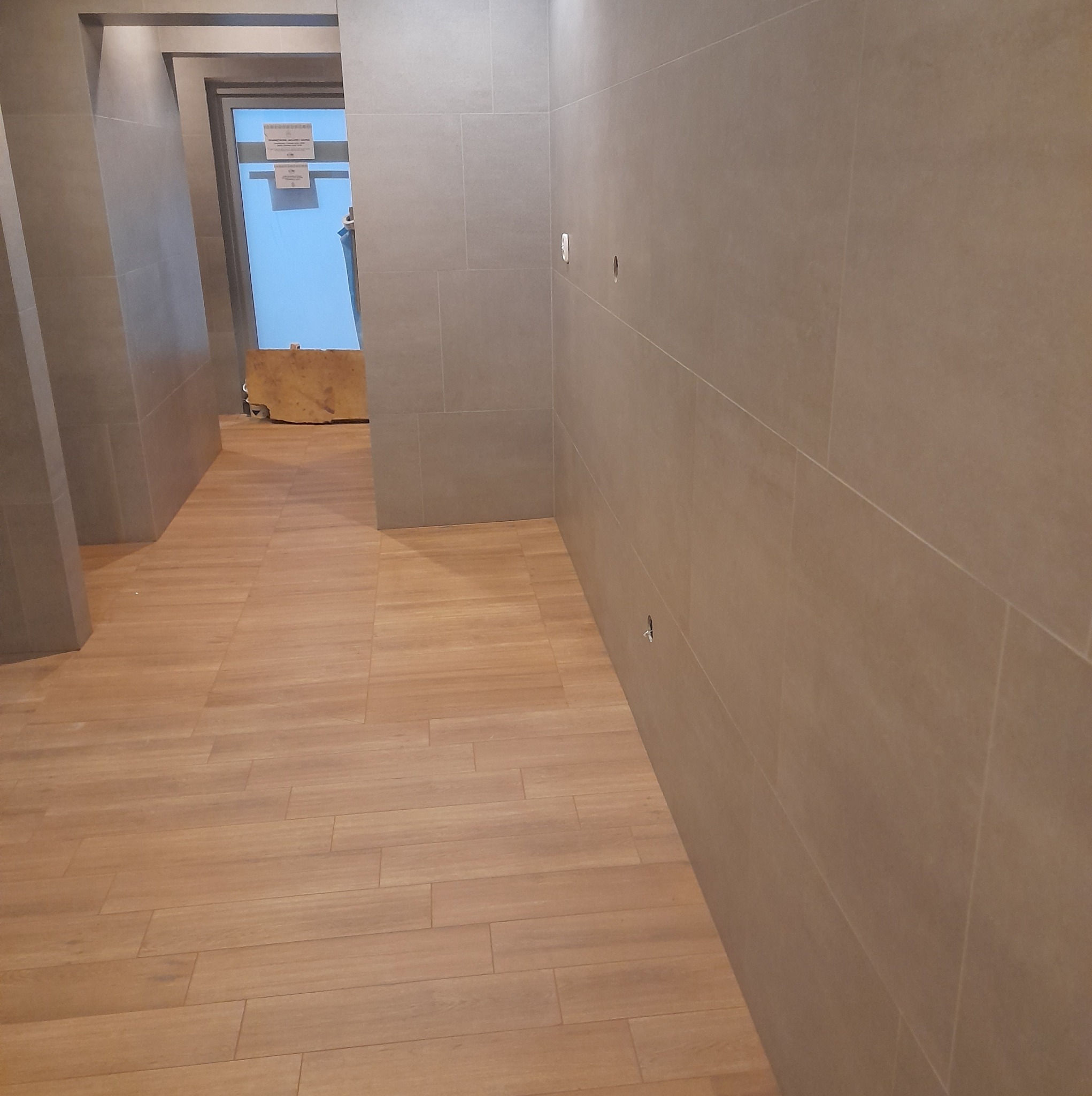 Korytarz wykończony płytkami ceramicznymi w odcieniach beżu i szarości, z widocznymi otworami na gniazdka elektryczne na ścianie i fragmentami dokumentów za szybą w tle.