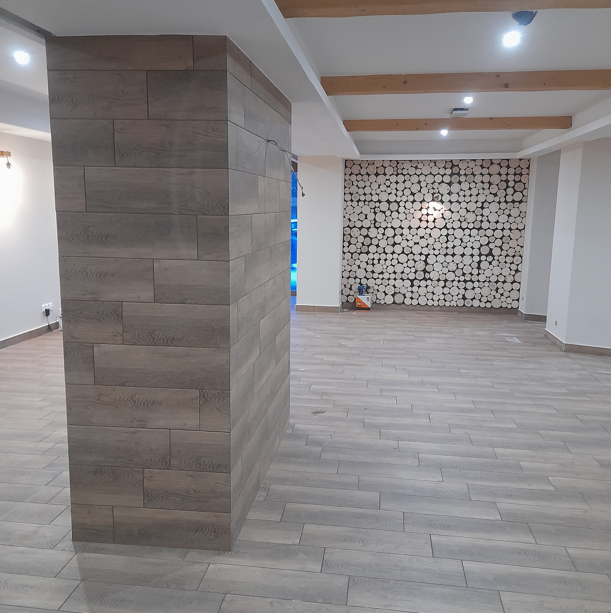 Wykończone wnętrze: podłoga i filar wyłożone panelami imitującymi drewno, dekoracyjna ściana z przekrojów pni drzew, drewniane belki stropowe, widoczne oświetlenie punktowe.