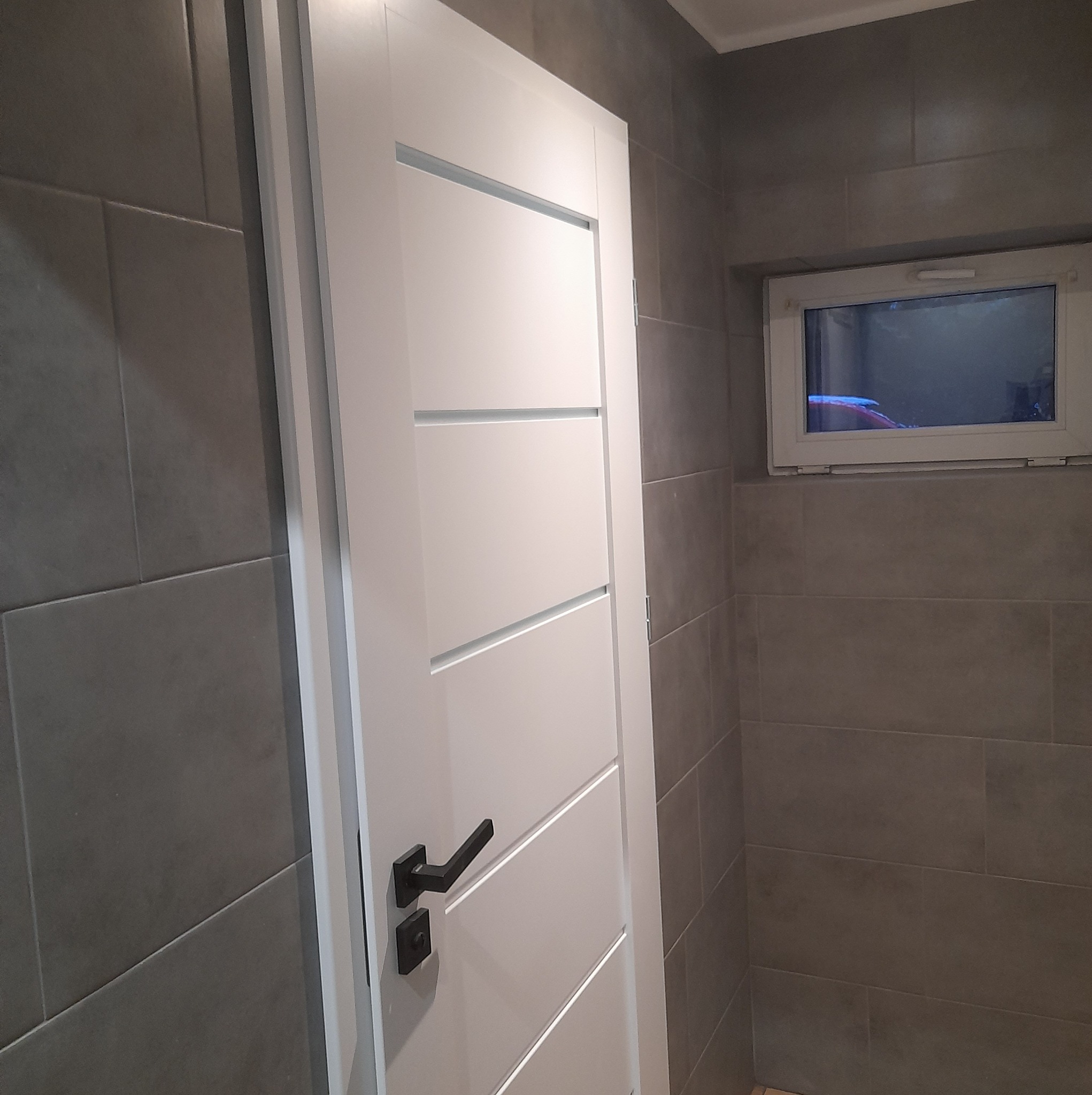 Białe drzwi wewnętrzne z poziomymi frezami i czarną klamką na tle szarych płytek w łazience z małym oknem.