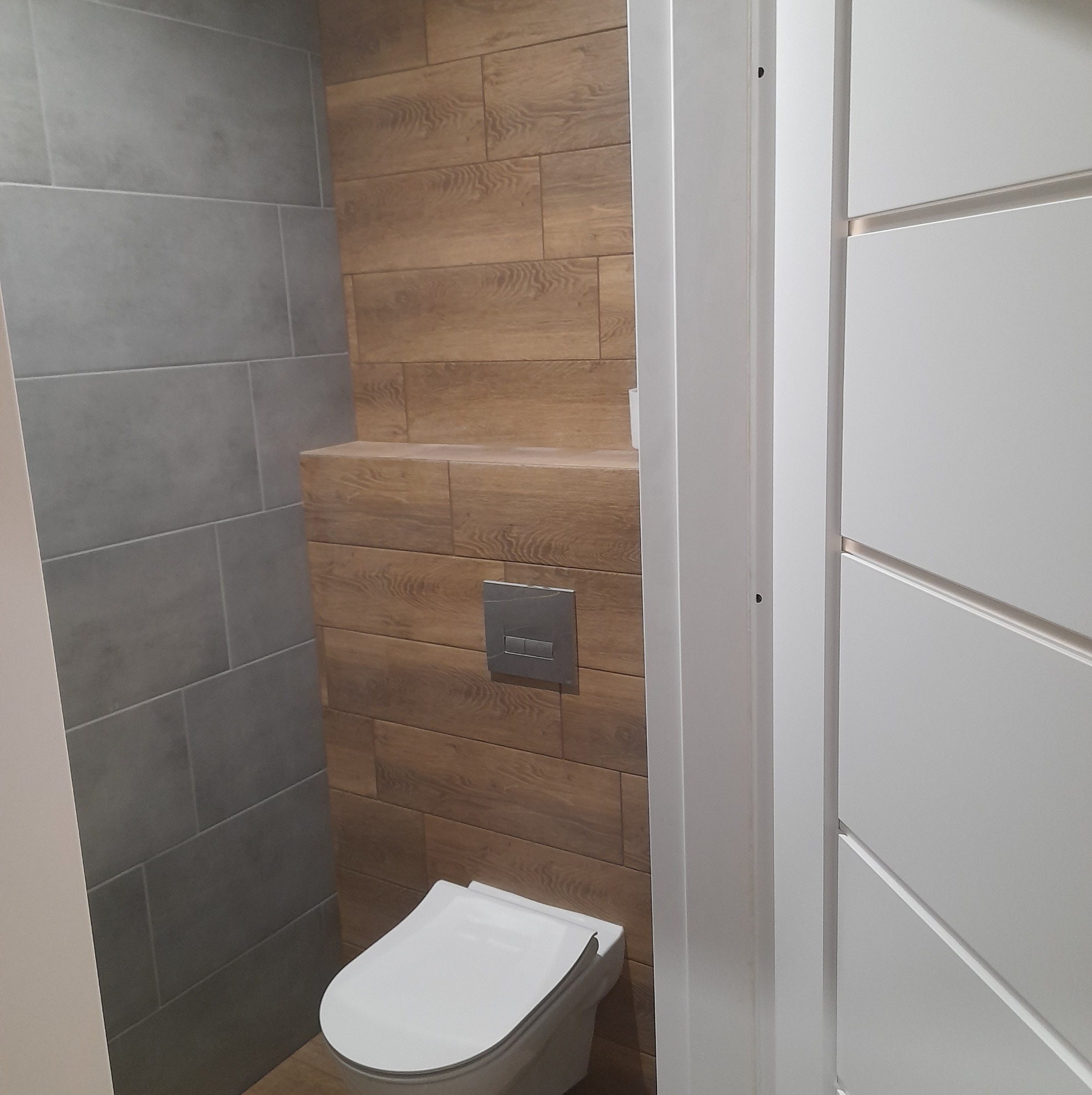 Nowoczesna toaleta podwieszana z szarymi i drewnopodobnymi płytkami, widoczny przycisk spłuczki, białe drzwi z frezowaniem.