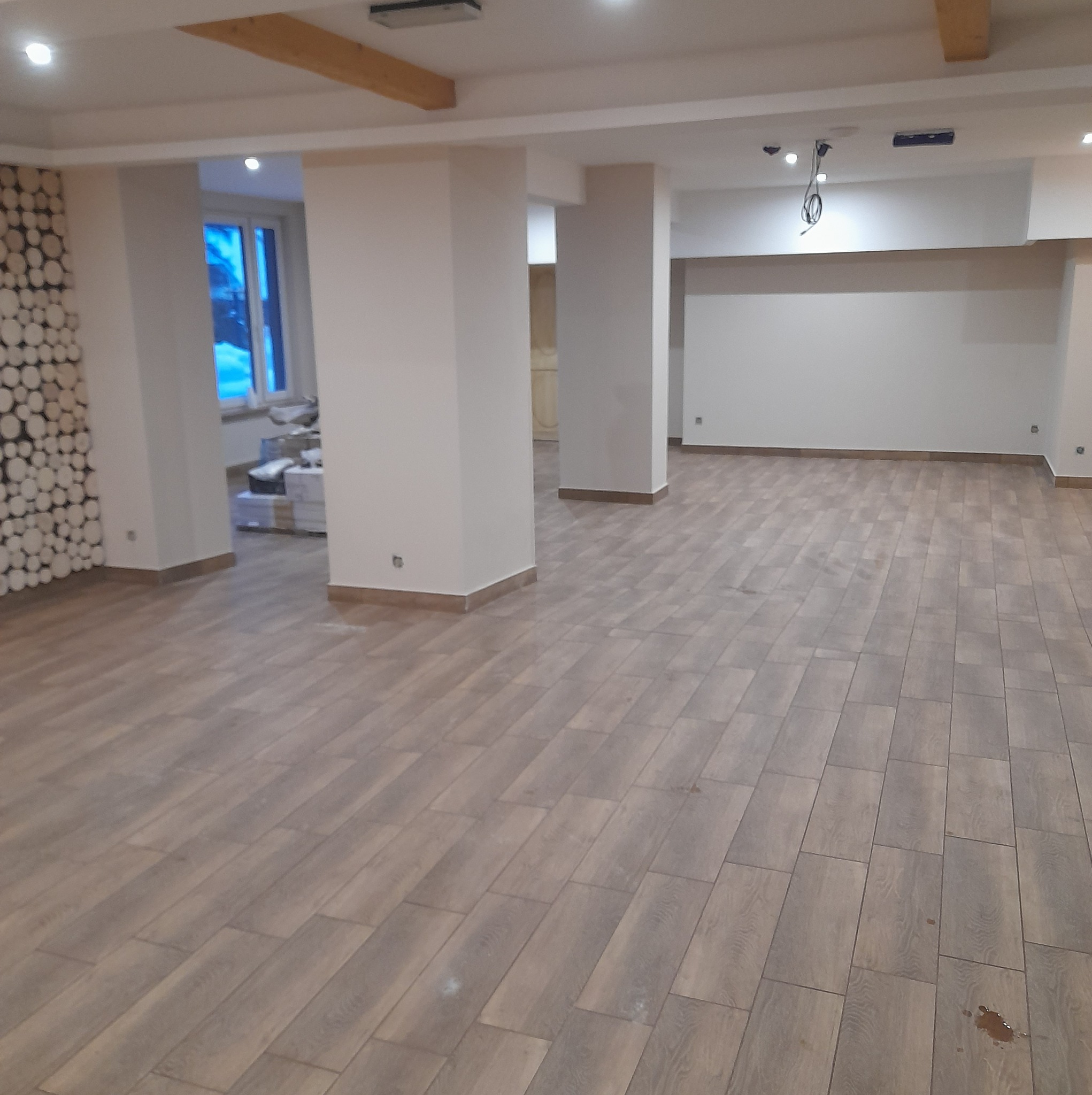 Przestronne wnętrze z ułożonymi panelami podłogowymi w odcieniach brązu, widoczne kolumny podtrzymujące strop i dekoracyjna ściana z drewnianych krążków, w tle okno z widokiem na zewnątrz.