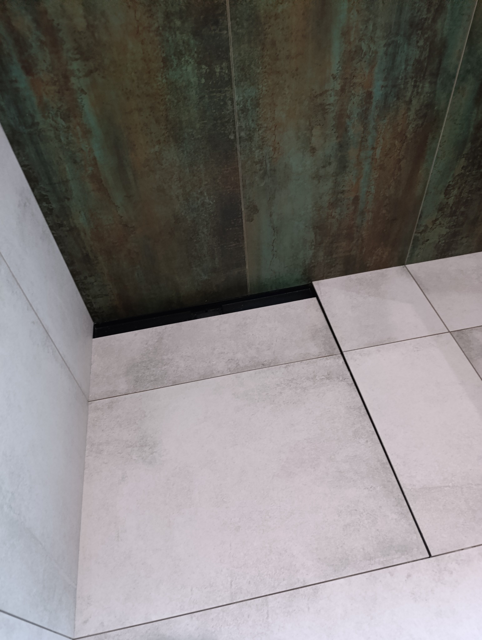 Połączenie płytek podłogowych w odcieniach szarości z płytkami ściennymi imitującymi rdzę, oddzielone czarną listwą wykończeniową.