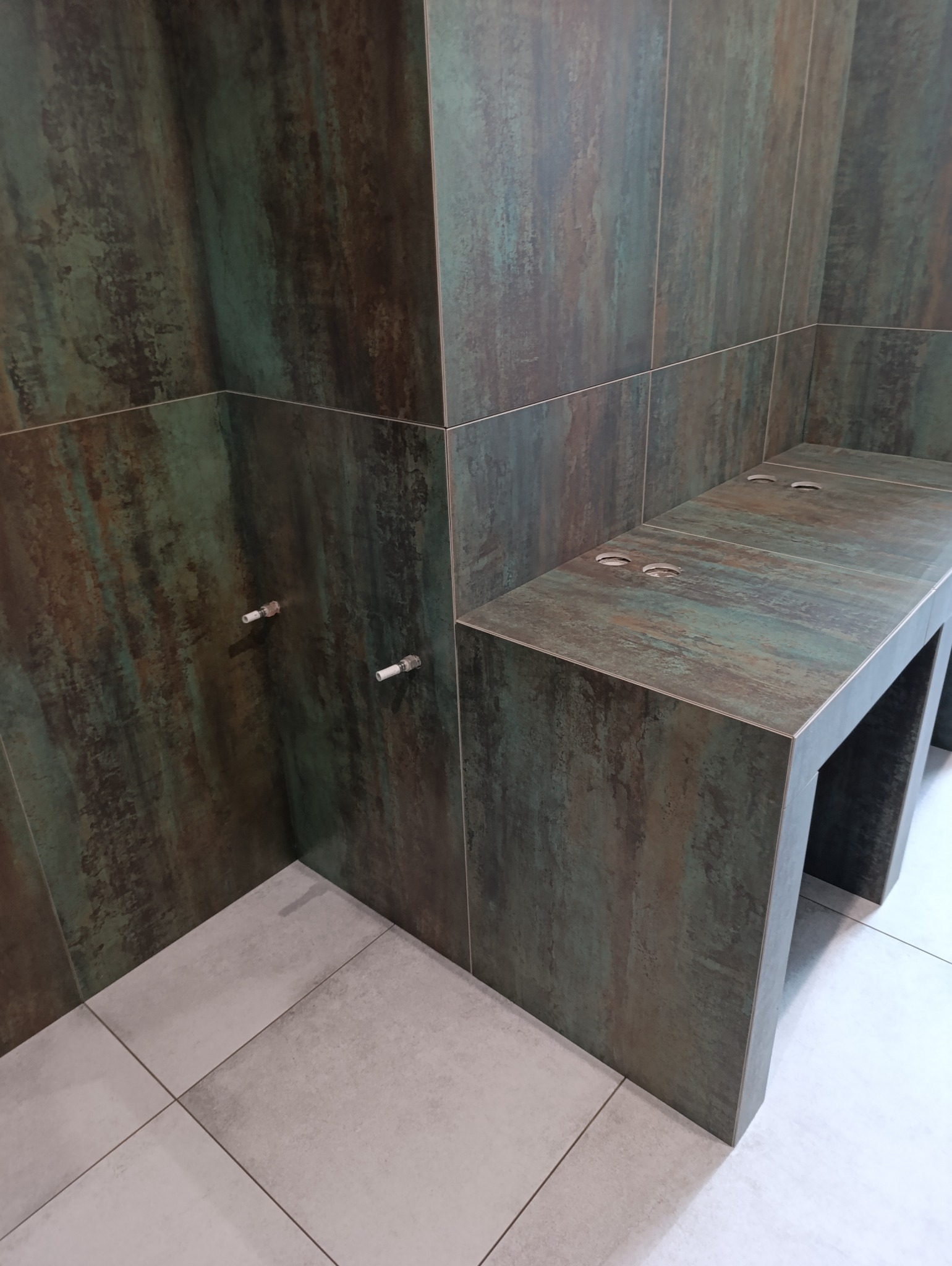 Wykończona kabina prysznicowa z siedziskiem, wyłożona płytkami imitującymi rdzę, z widocznymi przyłączami wodnymi i jasnymi płytkami podłogowymi.