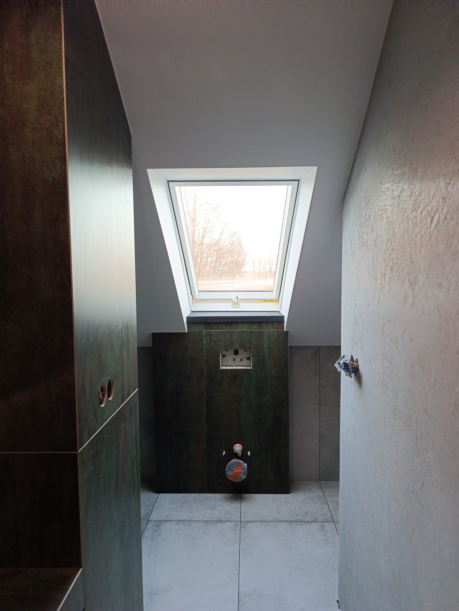 Wnętrze łazienki na poddaszu z oknem dachowym, widoczne płytki podłogowe w odcieniach szarości, ściana z dekoracyjną strukturą, zabudowa podtynkowego systemu WC z płytkami imitującymi rdzę...