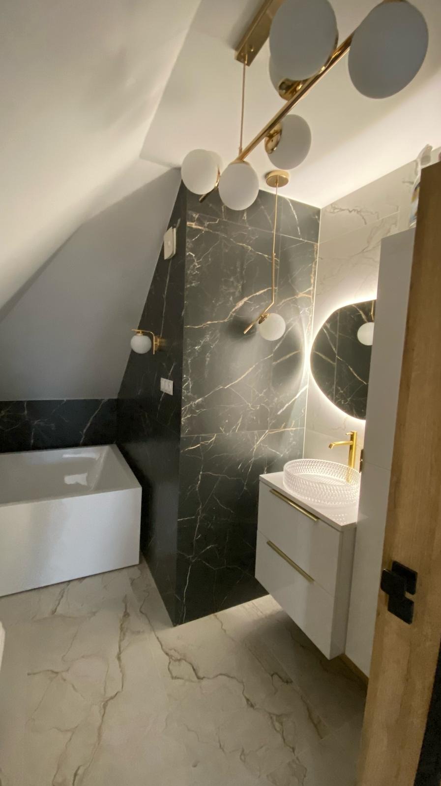 Nowoczesna łazienka na poddaszu z wanną, umywalką na szafce z złotymi uchwytami, okrągłym lustrem podświetlanym LED i czarnymi płytkami imitującymi marmur z złotymi żyłami.