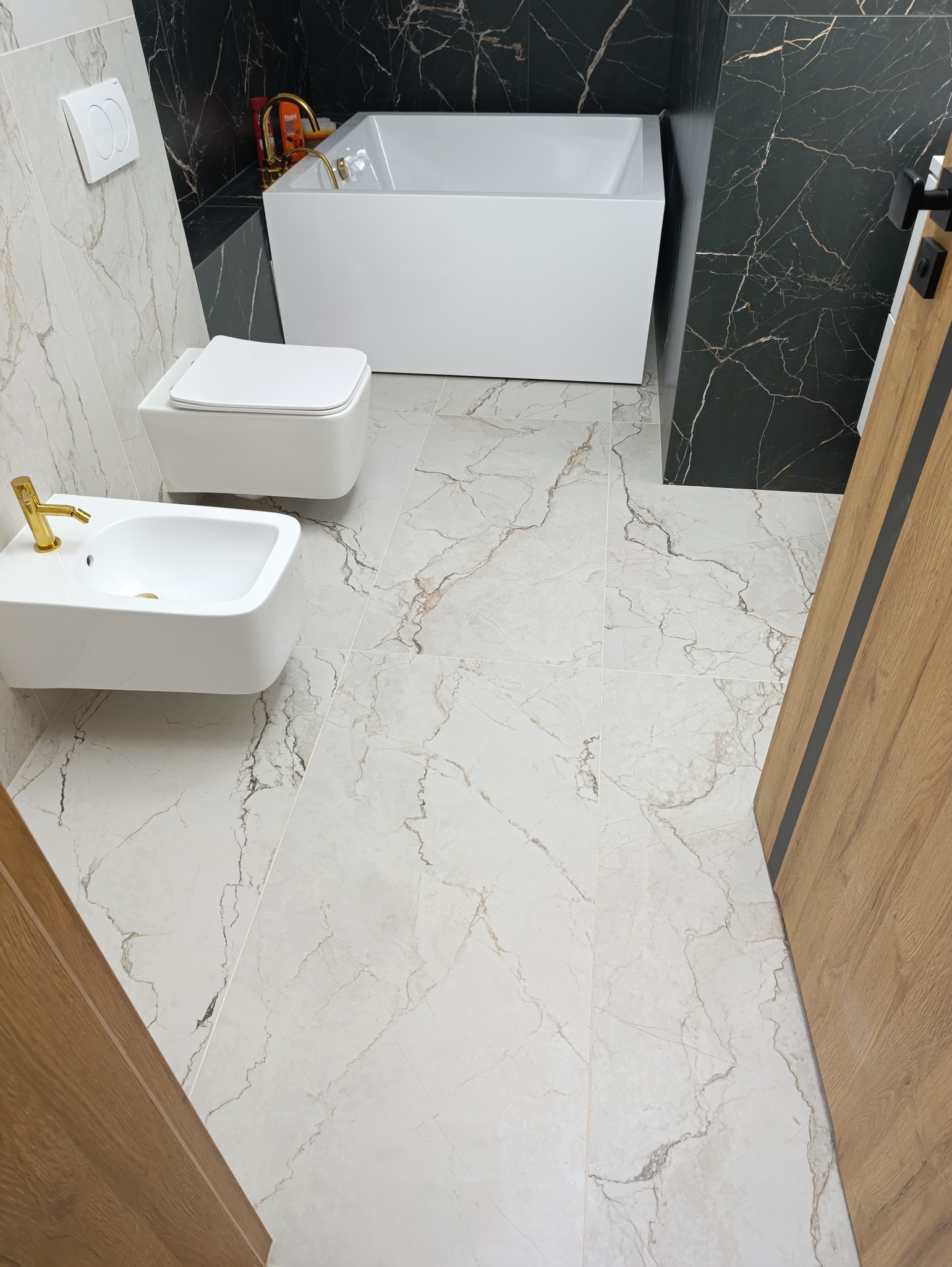 Nowoczesna łazienka z białą armaturą, złotą baterią i czarnymi płytkami imitującymi marmur na ścianach, jasne płytki podłogowe z marmurkowym wzorem, widok na wannę, toaletę i bidet.