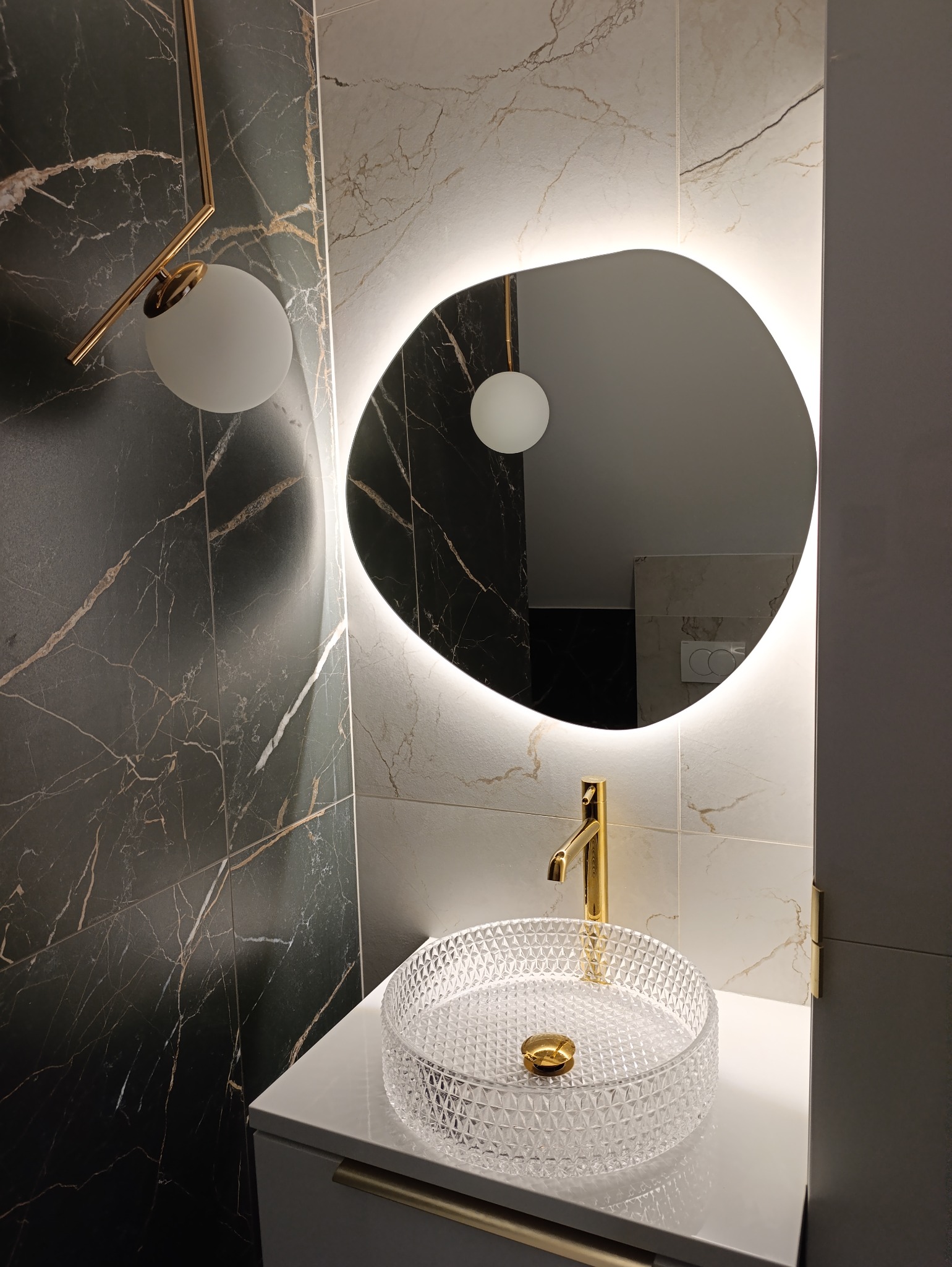 Nowoczesna łazienka z umywalką kryształową na blacie, złotą armaturą i podświetlanym lustrem o nieregularnym kształcie, ściany wyłożone płytkami imitującymi marmur w dwóch odcieniach.