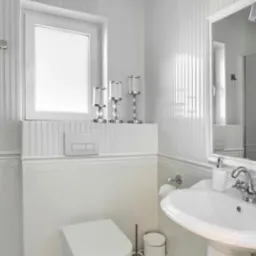 Elegancka, biała łazienka z toaletą podwieszaną, umywalką na postumencie i dekoracyjnymi świecznikami na parapecie okiennym.