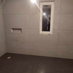 GRUCH - FACH Kewin Pałys - Wykończona łazienka w Konieczkowej: szare płytki ścienne, ciemna podłoga, okno z białymi ramami. Widoczne otwory na armaturę i wnęka w ścianie.