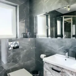 Elegancka łazienka z szarymi płytkami imitującymi marmur, białą armaturą i szafką pod umywalką z marmurowym wzorem. Widoczne oświetlenie sufitowe i duże lustro.