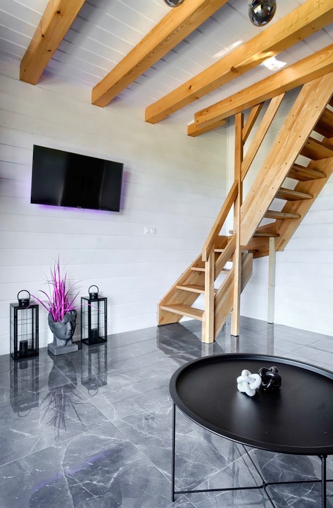 Jasne drewniane schody w nowoczesnym wnętrzu z białymi ścianami, widocznymi belkami stropowymi, szarą marmurową podłogą i czarnym stolikiem kawowym z dekoracjami.