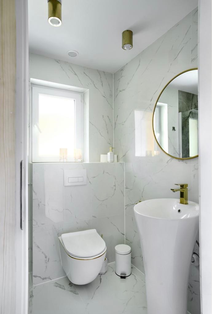 Elegancka, mała łazienka z białymi płytkami imitującymi marmur, złotymi akcentami w postaci baterii i ramy lustra, oraz wiszącą toaletą.