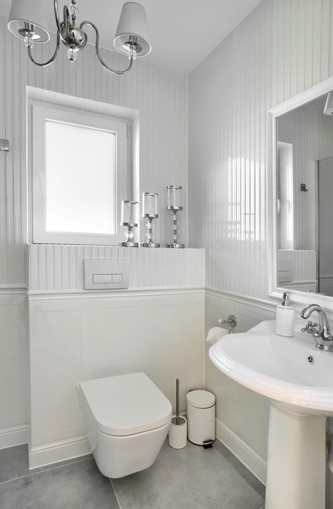 Elegancka, biała łazienka z toaletą podwieszaną, umywalką na postumencie i dekoracyjnymi świecznikami na parapecie okiennym.