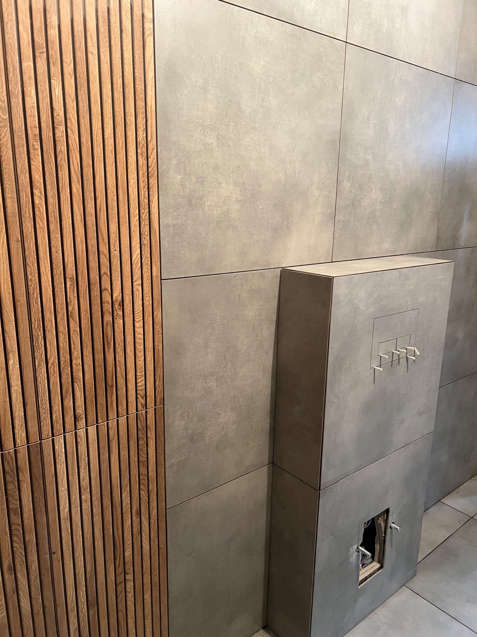 Szare, wielkoformatowe płytki na ścianie z drewnianą okładziną i zabudową stelaża WC z widocznymi elementami instalacji. Minimalistyczne wykończenie.