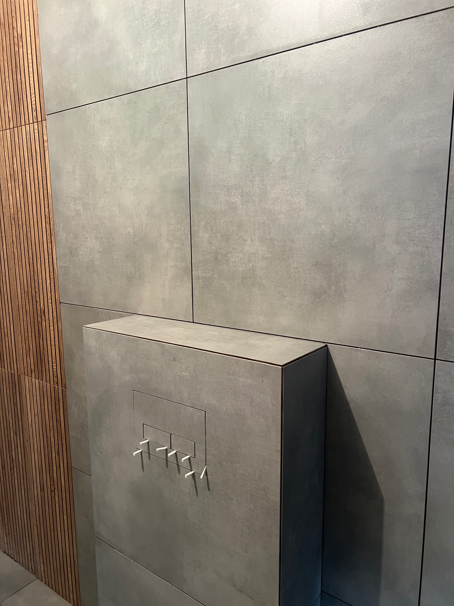 Minimalistyczna łazienka: szare, wielkoformatowe płytki imitujące beton, drewniane lamele i zabudowa stelaża WC z przyciskiem w kolorze płytek.