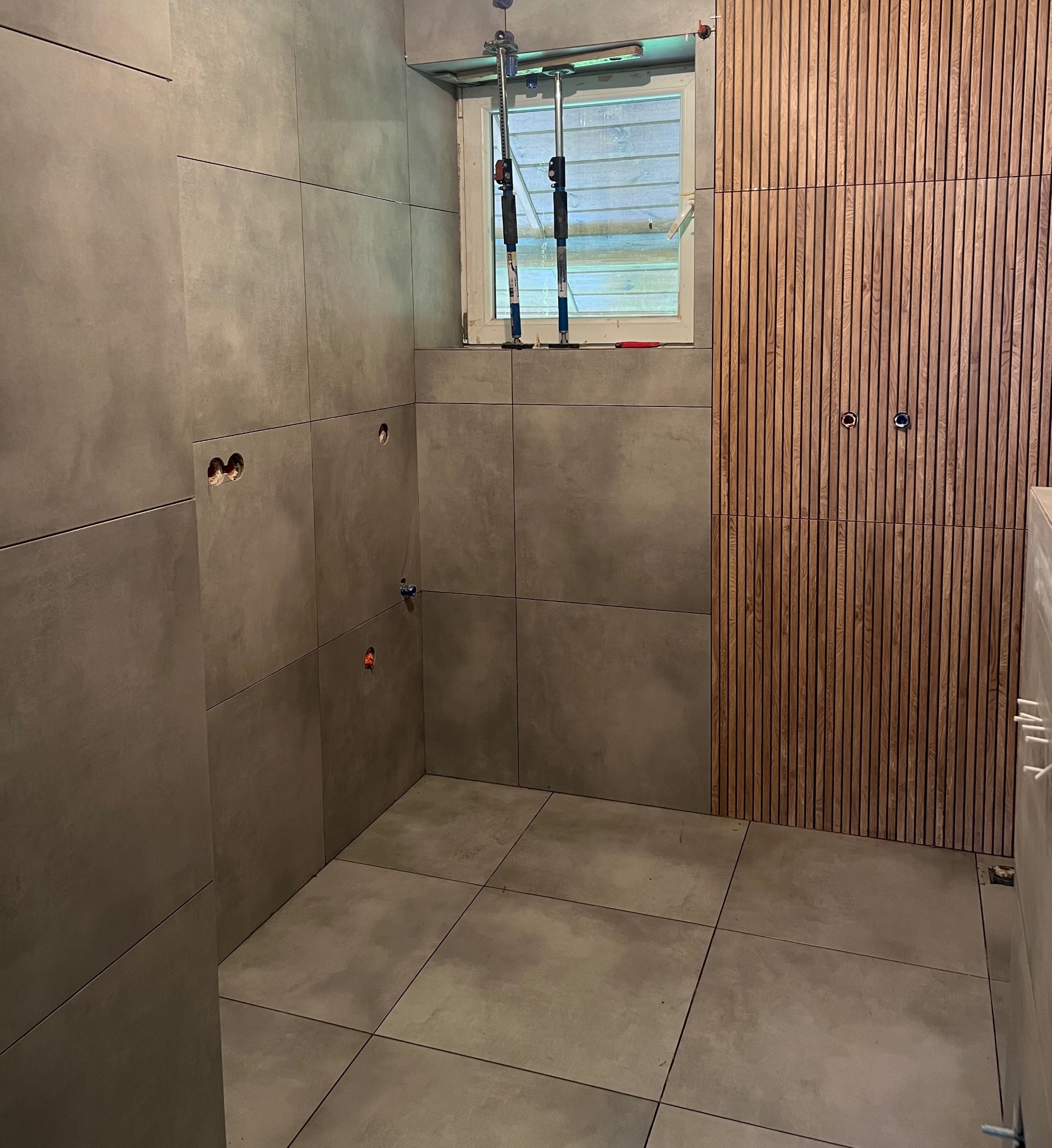 Wykończona łazienka z szarymi płytkami i drewnianą ścianą. Widoczne narzędzia glazurnicze przy oknie. Minimalistyczny design z elementami naturalnego drewna.