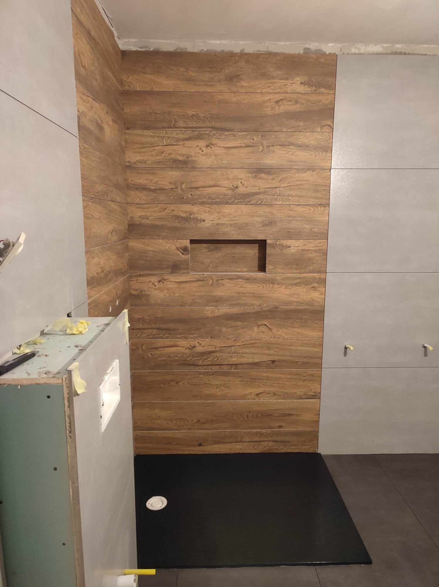 Wykończona strefa prysznicowa z płytkami imitującymi drewno i szarymi, dużymi płytami. Widoczny czarny brodzik i fragment zabudowy pod umywalkę.