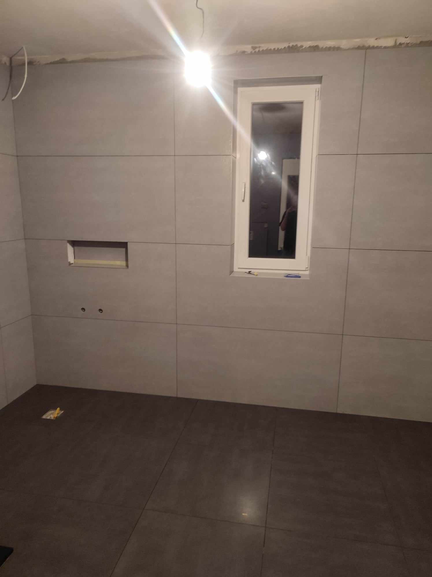 Wykończona łazienka w Konieczkowej: szare płytki ścienne, ciemna podłoga, okno z białymi ramami. Widoczne otwory na armaturę i wnęka w ścianie.