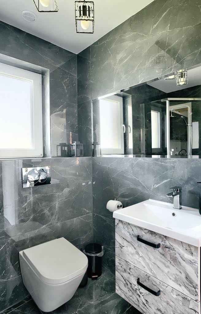 Elegancka łazienka z szarymi płytkami imitującymi marmur, białą armaturą i szafką pod umywalką z marmurowym wzorem. Widoczne oświetlenie sufitowe i duże lustro.