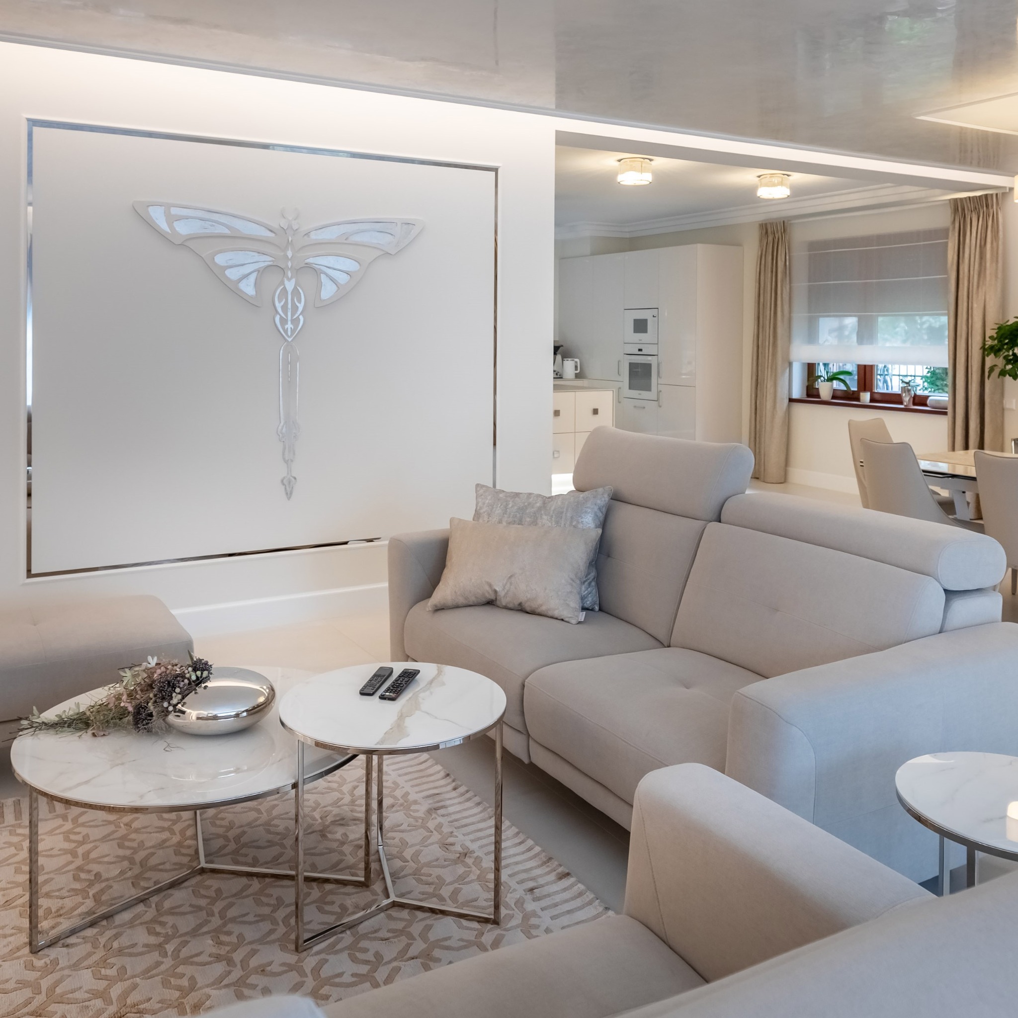 Jasny salon z beżową sofą, stolikami z marmurowymi blatami i dekoracją ścienną w kształcie motyla, widok na kuchnię w tle.