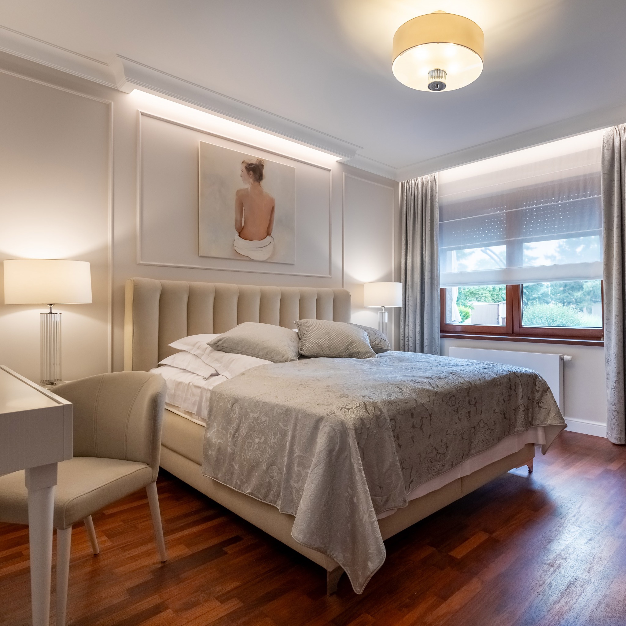 Elegancka sypialnia w Krzyszkowie z drewnianą podłogą, beżowym łóżkiem z pikowanym zagłówkiem, obrazem kobiety na ścianie i lampami nocnymi z kryształowymi podstawami.